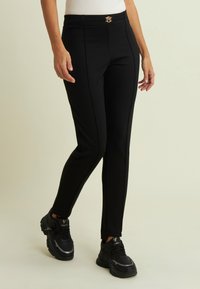 Pantaloni neri aderenti realizzati in tessuto elastico. Presentano una fibbia del cinturone tono oro e cuciture decorative lungo la parte anteriore. Indossati con scarpe da ginnastica nere.