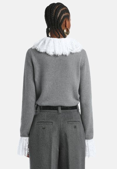 Pilkas megztinis su baltu nėriniu apykakle. Rankovės dekoruotos nėriniu, derinamos su tekstūruotomis pilkomis kelnėmis ir juodu diržu.