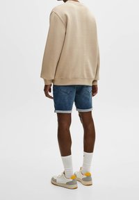 Sweat-shirt beige à manches longues, associé à un short en jean bleu, et des baskets neutres avec des accents jaunes. Fond blanc uni.