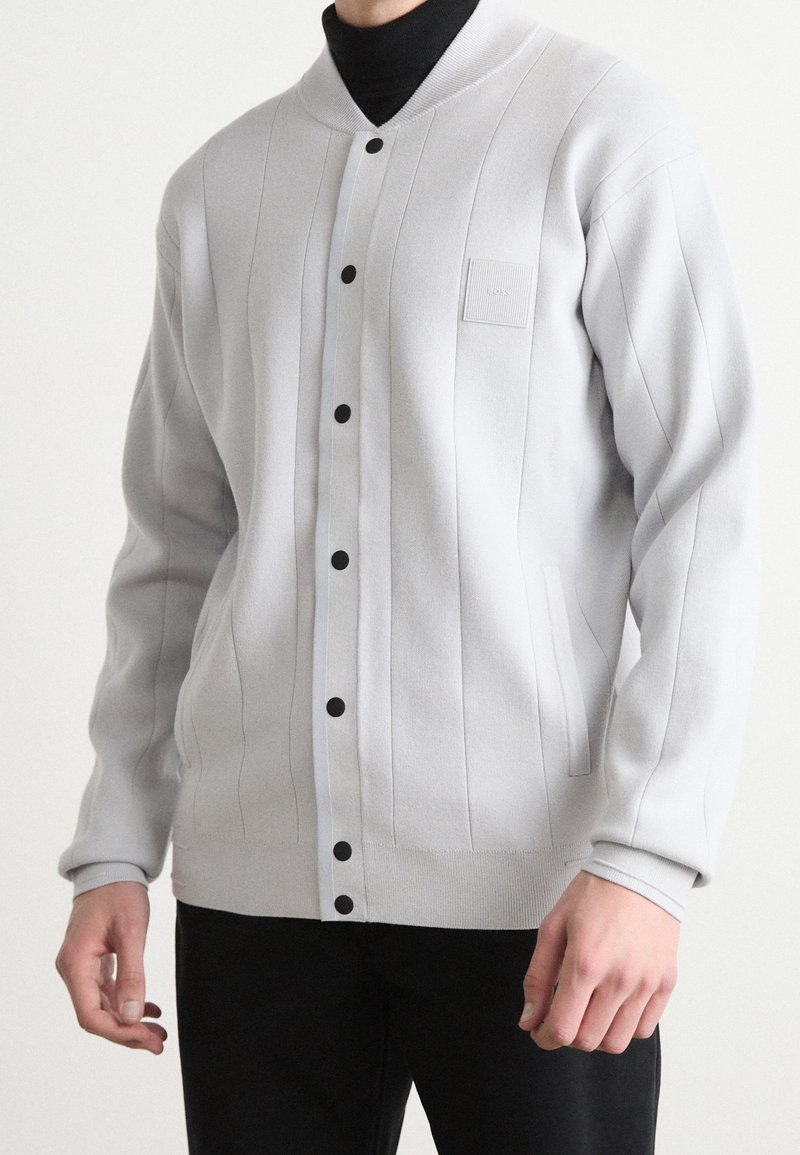 Cardigan gris clair boutonné avec côtes verticales, patch logo sur la poitrine, porté sur un col roulé noir avec un pantalon noir.