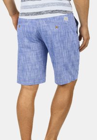 Blå linneshorts med strukturert overflate, to baklommer med knappelukking og et merke på linningen. Over kneet lengde.