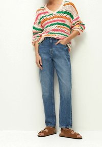 Gestreifter Strickpullover in Pink, Grün, Orange und Gelb, kombiniert mit blauen, geraden Jeans und braunen Sandalen mit goldenen Akzenten.