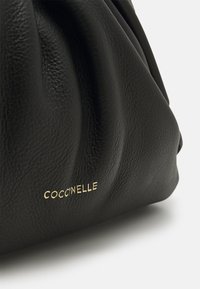 Black leather handbag with gathered design, featuring a gold "Coccinelle" logo. Smooth texture and medium-size, highlighting soft folds.