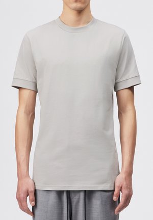 Man wearing plain light gray short-sleeve T-shirt with ribbed cuffs and neck, paired with loose gray pants against white background.