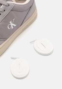 Szürke sneaker bézs talppal, sima anyagból, dombornyomott logóval. Két fehér, kerek cipőfűző látható a cipő mellett.