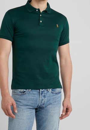 Man draagt een donkergroen polohemd met korte mouwen en twee knopen, gecombineerd met lichtblauwe spijkerbroek, voor een effen achtergrond.