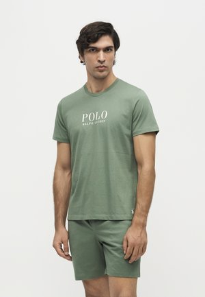 Млад мъж, носещ бяла тениска Polo Ralph Lauren в маслиненозелен цвят и съвпадащи шорти, стои пред обикновен бял фон.