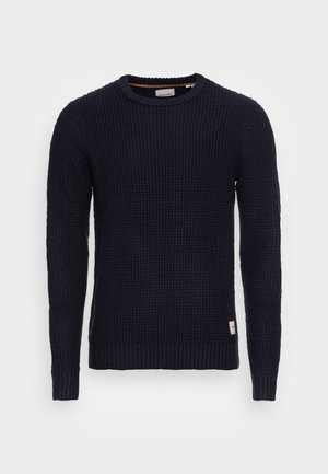 Pull en tricot bleu marine avec un motif texturé, col rond, et poignets et ourlet côtelés. Il est orné d'une petite étiquette en bas à gauche.