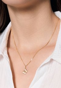 Złoty wisiorek w kształcie motyla na cienkim łańcuszku, z wyraźnym wzorem i błyszczącymi akcentami. Noszony na białej koszuli z kołnierzykiem.