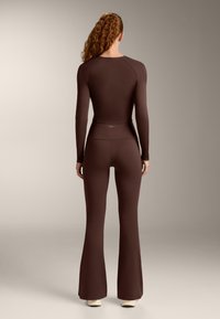 Vrouw staand met de rug naar de camera, gekleed in een donkerbruin nauwsluitend shirt met lange mouwen en uitlopende broek, met crèmekleurige schoenen, tegen een neutrale achtergrond.