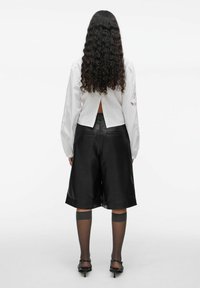 Femme avec de longs cheveux bouclés portant un chemisier blanc à manches longues, un short en cuir noir jusqu'aux genoux, des chaussettes hautes en voile jusqu'aux genoux et des talons noirs.