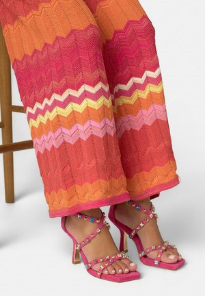 Legs wearing multicolored zigzag knit pants and pink high-heeled sandals decorated with colorful gemstones, resting near wooden chair legs.