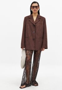 Blazer oversize in finta pelle scamosciata marrone con chiusura a bottone e tasche a patta, abbinato a pantaloni ampi inpizzo neri con un motivo floreale.