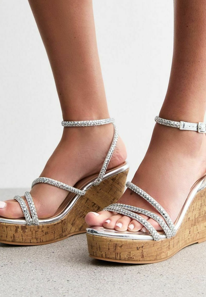 Sandalias de cuña plateadas con tiras entrelazadas que presentan un tacón de corcho, tiras texturizadas con strass y una base metálica brillante.