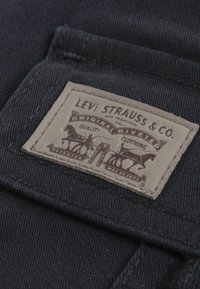 Etichetă din piele maro cu text și imagini în relief, cusută pe material denim albastru închis, afișând logo-ul și detaliile Levi Strauss.