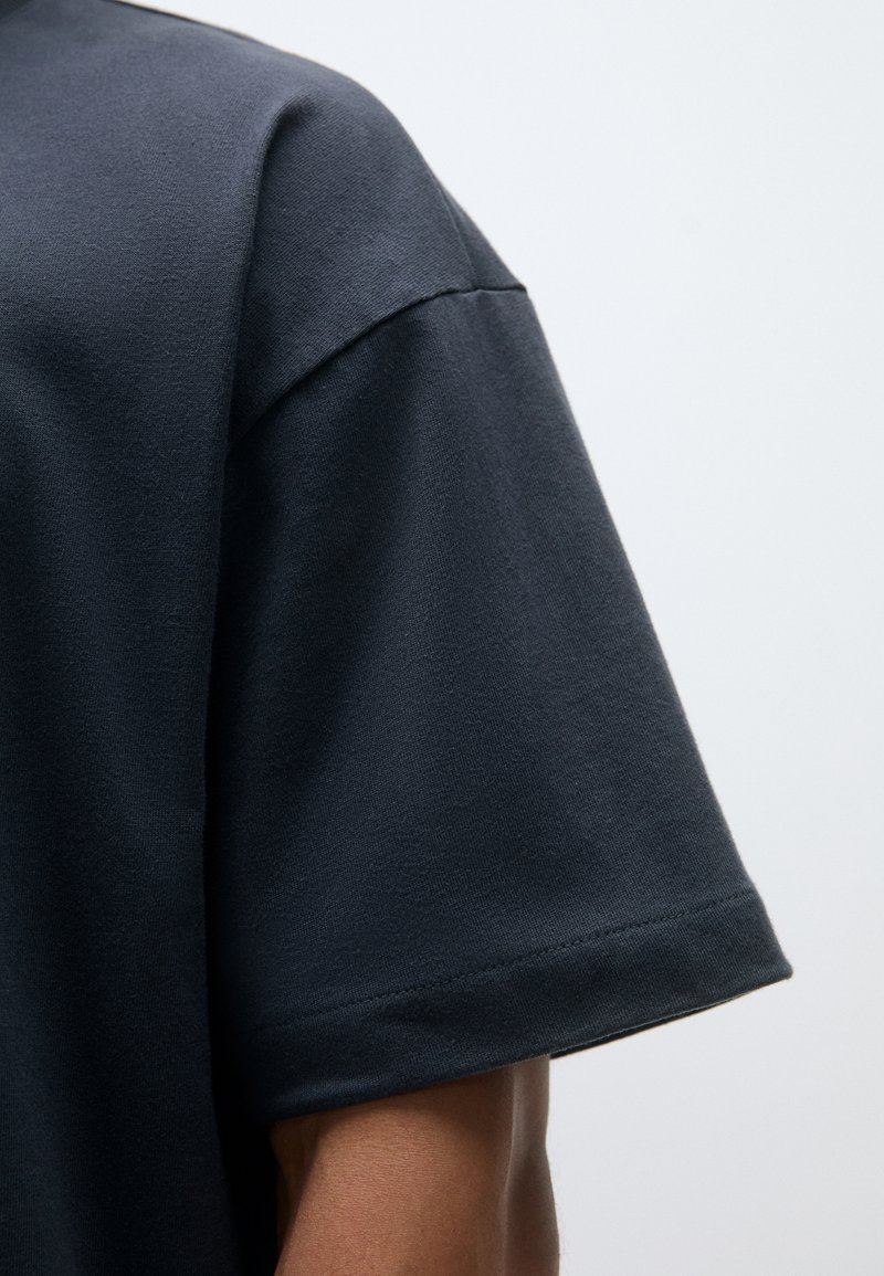 T-shirt a maniche corte grigio scuro realizzato in morbido cotone. Presenta un collo a giro e una vestibilità rilassata, con cuciture visibili sulle maniche.