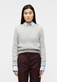 Γυναίκα με μακριά σκούρα μαλλιά που φοράει ανοιχτό γκρι πουλόβερ πάνω από μπλε πουκάμισο με γιακά και σκούρο βυσσινί παντελόνι, στέκεται μπροστά σε απλό λευκό φόντο.