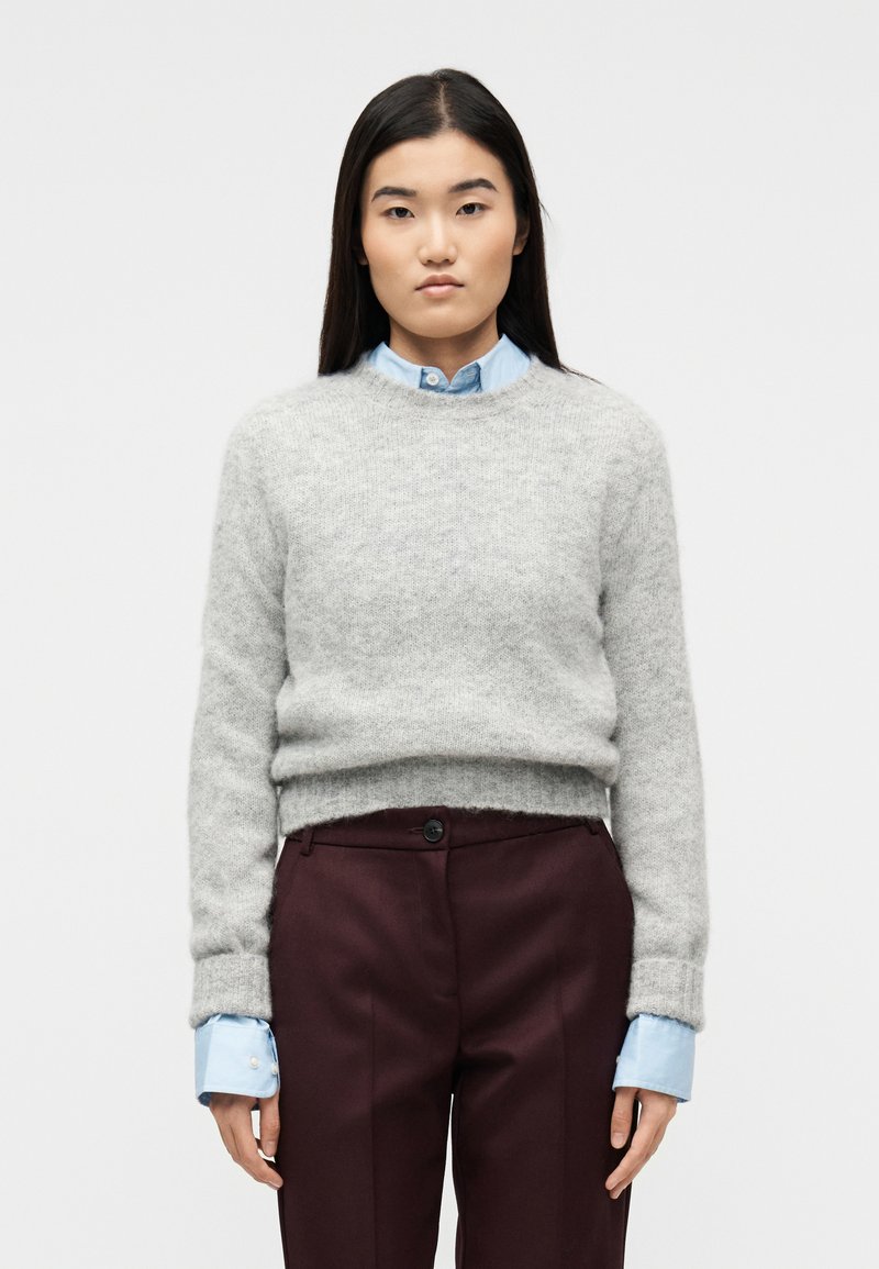 Γυναίκα με μακριά σκούρα μαλλιά που φοράει ανοιχτό γκρι πουλόβερ πάνω από μπλε πουκάμισο με γιακά και σκούρο βυσσινί παντελόνι, στέκεται μπροστά σε απλό λευκό φόντο.