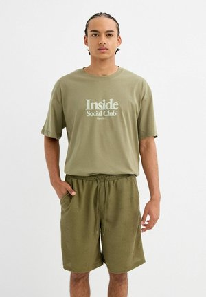 Joven con el cabello trenzado, lleva una camiseta verde olivo con la inscripción "Inside Social Club" y pantalones cortos texturizados a juego, de pie con una mano en el bolsillo.
