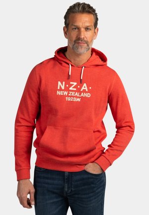Man met grijze baard, gekleed in een rode hoodie met de tekst "N.Z.A. New Zealand 1923" en een blauwe spijkerbroek, staand tegen een effen lichte achtergrond.