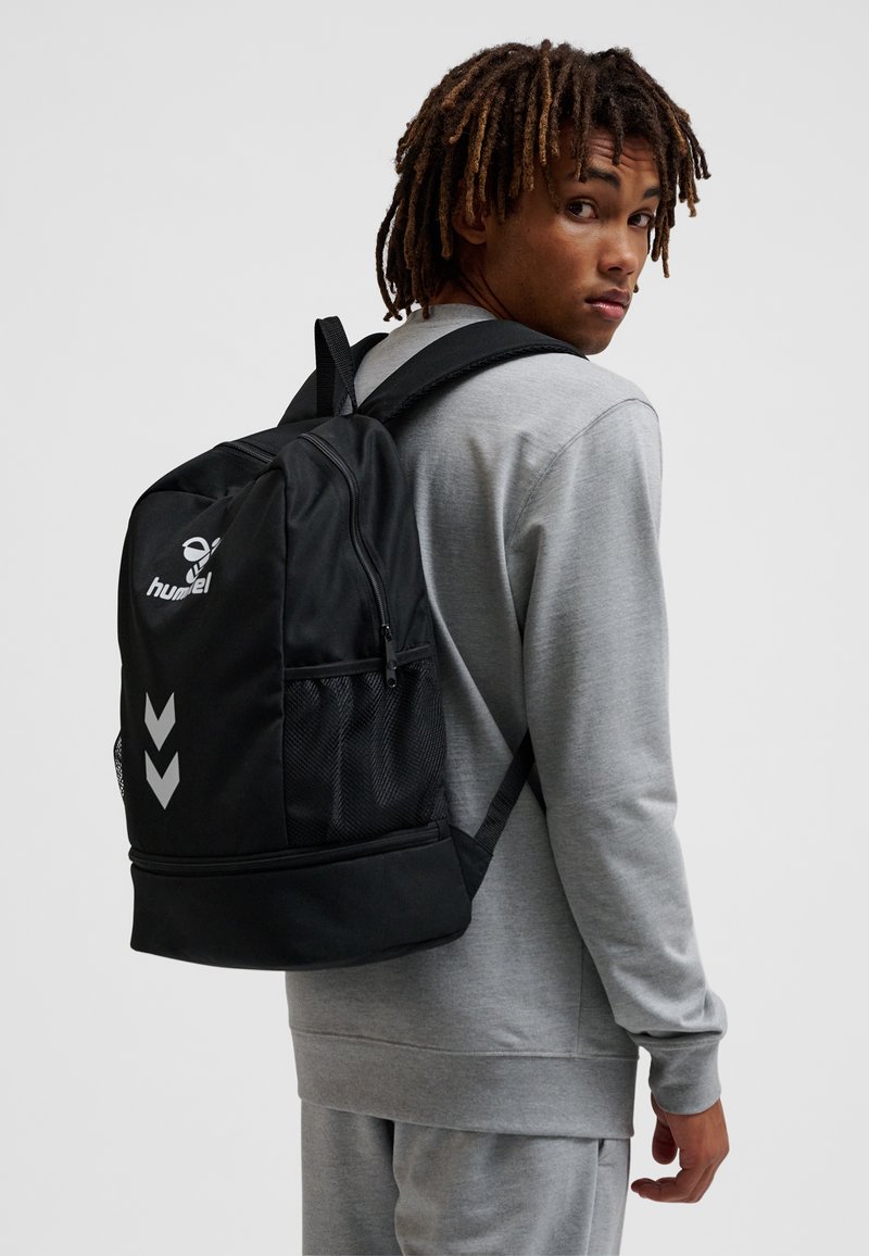 Jong persoon met dreadlocks, gekleed in een grijs trainingspak en een zwarte rugzak met wit logo en chevronpatroon, kijkt over zijn schouder.