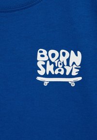 Blå bomullströja med en vit grafisk design som har texten "FÖDD FÖR ATT ÅKA" och en skateboard-illustration nedanför, designad med rundade bokstäver.