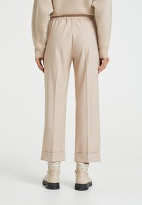 Beige mukautetut housut rentoilla leikkauksilla, joiden lahkeet ovat käännetyt ja vyötäröllä on joustava vyö, yhdistettynä beigeihin nilkkabootseihin ja tiukkoihin vaaleisiin sukkiin.