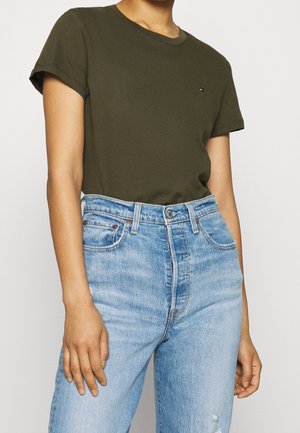 Persona con camiseta de manga corta color verde oliva metida dentro de unos vaqueros de mezclilla de tiro alto azul claro, con las manos relajadas a los lados.