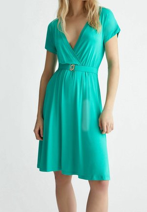 Kobieta w turkusowej sukience kopertowej z krótkimi rękawami i pasem w tym samym kolorze z złotą klamrą, stojąca na jednolitym tle.