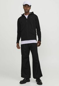 Schwarze Zip-Up Kapuzenjacke, kombiniert mit lockeren schwarzen Hosen. Das Modell trägt eine weiße Mütze und geschichtete Kleidung. Schwarze Stiefel runden den Look ab.