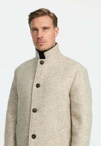 Béžový kabát z vlnenej zmesi s vysokým golierom, gombíkovým zapínaním a textúrovaným vzorom. Má dve bočné vrecká a štíhly strih.