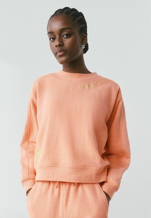 Femme noire aux cheveux tressés portant un sweat ample couleur pêche et un pantalon assorti, debout devant un fond clair uni.