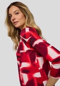 Blonde Frau, die ein langärmliges Oberteil mit abstraktem Muster in Rot, Weiß und Schwarz trägt und über ihre Schulter blickt vor einem einfarbigen Hintergrund.