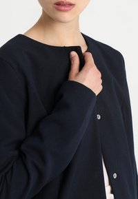Mujer con un abrigo azul marino sosteniendo el cuello cerca de su cuello, mostrando botones a presión y parte de una blusa de color claro debajo.