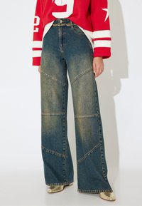 Vaqueros de mezclilla de pierna ancha y cintura alta con un lavado azul desgastado, que presentan costuras de panel en contraste y dobladillos sin rematar. Llevados con un sudadera roja.