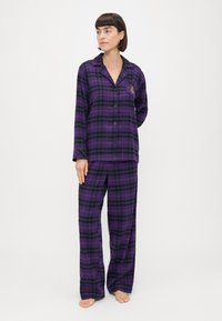 Πυτζάμα σε μωβ και μαύρη καρώ εκτύπωση, περιλαμβάνει πουκάμισο με κουμπιά και μακριά μανίκια καθώς και αντίστοιχα ελαφρώς φαρδιά παντελόνια. Εμπριμέ λεπτομέρεια στο πουκάμισο.