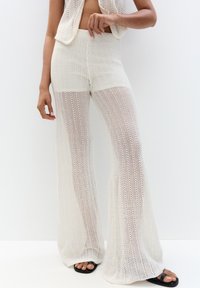 Witte gebreide twee-delige outfit met een mouwloze top en flared broek met een open geweven ontwerp en textuurstof. Zwarte sandalen getoond.