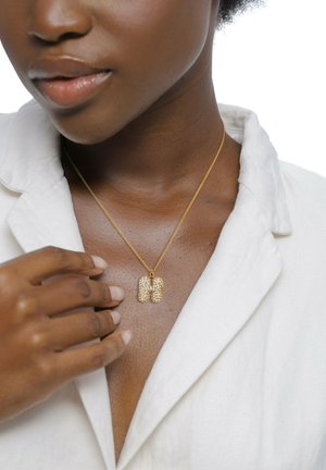 Goldkette mit einem strukturierten Anhänger, der ein Muster aus kleinen klaren Steinen zeigt. Das Modell trägt ein helles Hemd, die Hand ruht am Halsausschnitt.