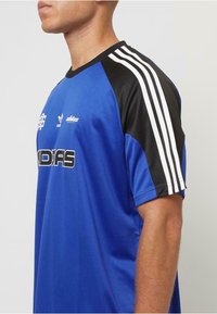 Blå idrottströja med svarta korta ärmar, tre vita ränder på axlarna och en grafisk logotyp på framsidan. Mjuk, andningsbar tyg.