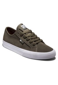 Zapatilla de lona verde olivo con suela de goma blanca, diseño con cordones, cuello acolchado y logo negro en la lengüeta. Detalle de superficie texturizada.