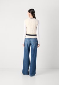 Beige Strickweste mit Zopfmuster, navyblau-gelb gestreiftem Saum, kombiniert mit hellblauen weit geschnittenen Jeans. Einfarbiger Hintergrund, Modell hat sich abgewandt.