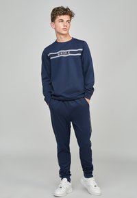 Conjunto de sudadera y joggers en color navy, fabricado con un material suave. Presenta un logo blanco y franjas en el pecho y los lados. Combinado con zapatillas blancas.
