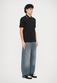 Camisa polo negra con logo estampado sutil, mangas cortas y detalles en el cuello blanco, combinada con jeans de pierna ancha y desgastados en azul y zapatos negros.