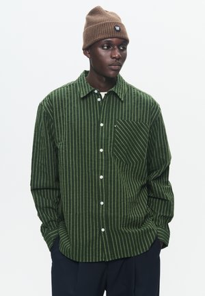 Green corduroy shirt with vertical stripes, button-up front, single chest pocket, and long sleeves. Worn with a brown ribbed beanie.