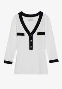 Cardigan bianco con accenti neri, scollo a V, chiusura frontale con bottoni decorati da anelli argentati e due dettagli di tasche nere sul petto.