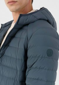 Marineblauwe gewatteerde jas met capuchon, voorzien van gestikte secties, een gladde textuur en een ronde logo patch op de linkermouw.
