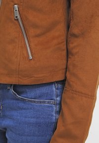 Bruine suède jas met zilveren ritsdetails, voorzien van een getailleerd ontwerp en een gladde textuur, gecombineerd met blauwe denim jeans.