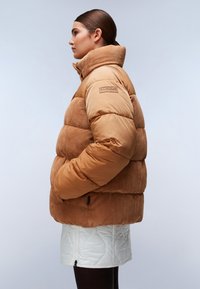 Kobieta w profilu, ubrana w mikstaną kurtkę z tanowego sztruksu i pikowanej puchówki z wysokim kołnierzem, białą pikowaną spódnicę i ciemne legginsy na szarym tle.