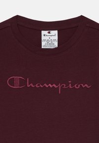 Tamsiai raudonas medvilninis marškinėliai su apvaliu iškirpimu ir siuvinėtu "Champion" logotipu šiek tiek šviesesnėje raudonoje atspalvyje.