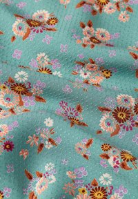Gemusterter Stoff mit einem türkisfarbenen Hintergrund und floralen Motiven in Orange, Rosa und Lila. Die Textur ist leicht strukturiert mit kleinen Punkten.